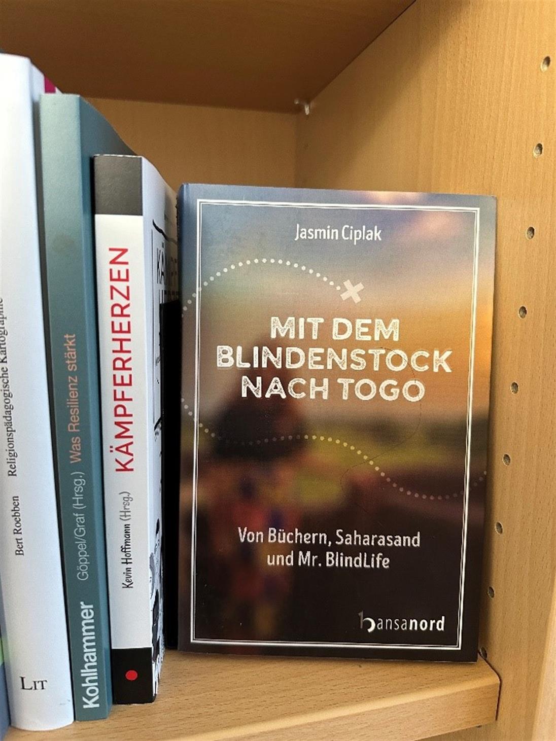 Bildbeschreibung: Das Bild zeigt die Titelseite des Buches: „Mit dem Blindenstock nach Togo – von Büchern, Saharasand und Mr. BlindLife“ in einem Bücherregal. 